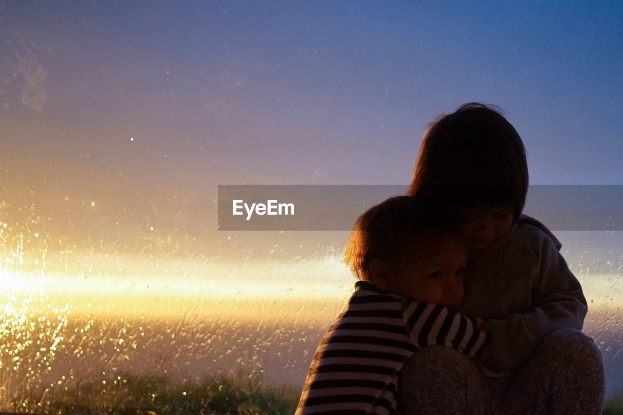 Siblings embracing against glass window during sunset