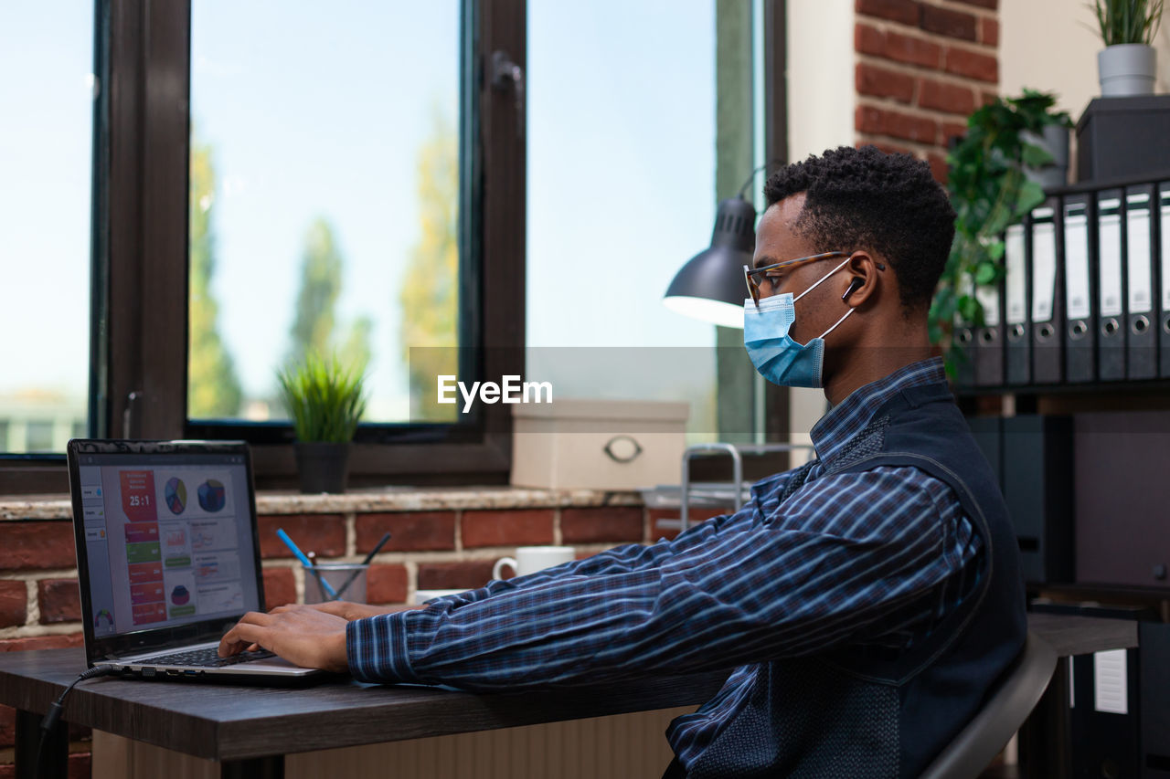 Side view of businessman wearing mask using laptop at office