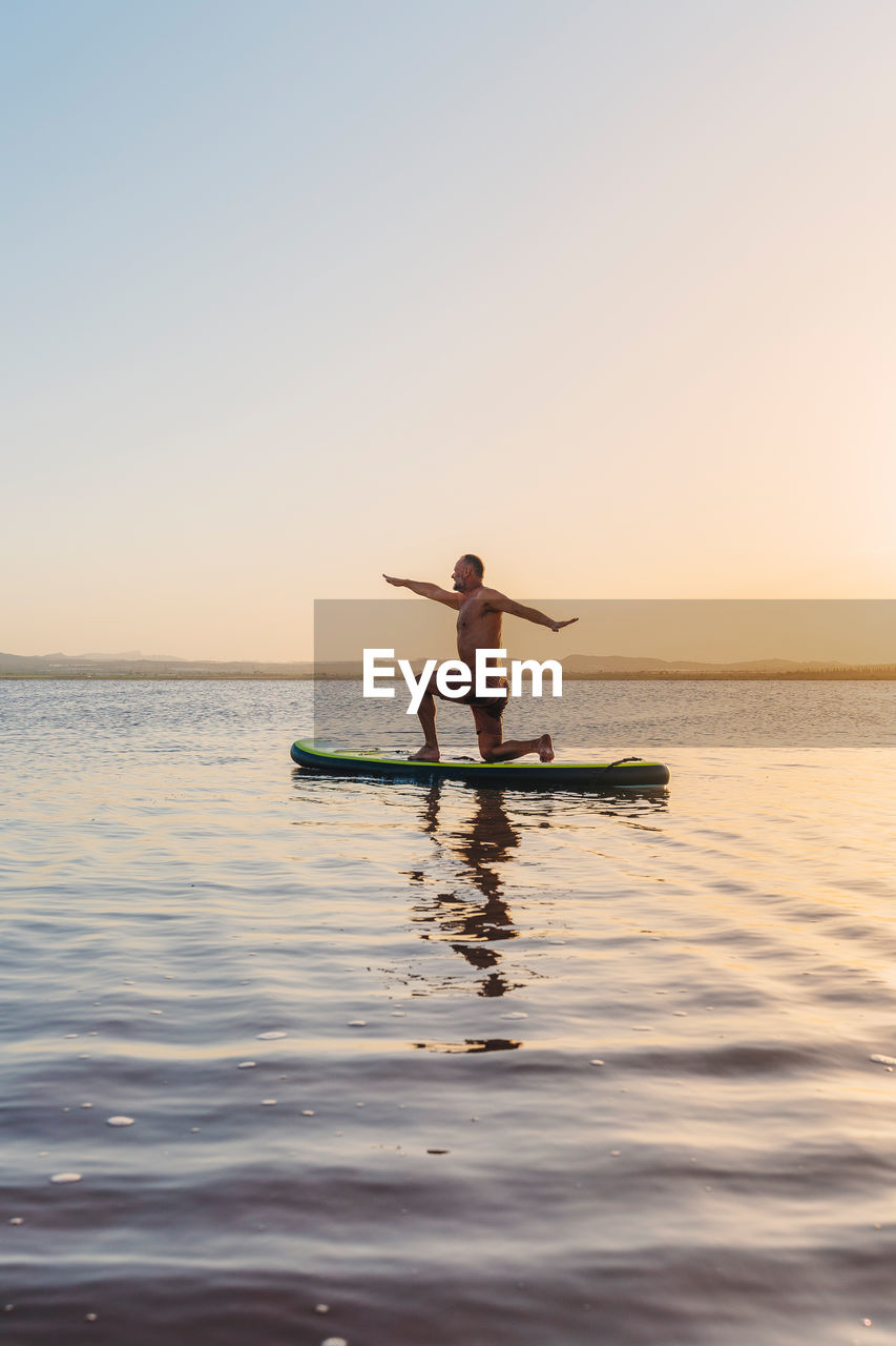 Full body side view of unrecognizable man doing variation of crescent lunge on knee asana while floating on paddle board in calm lake water during sunset