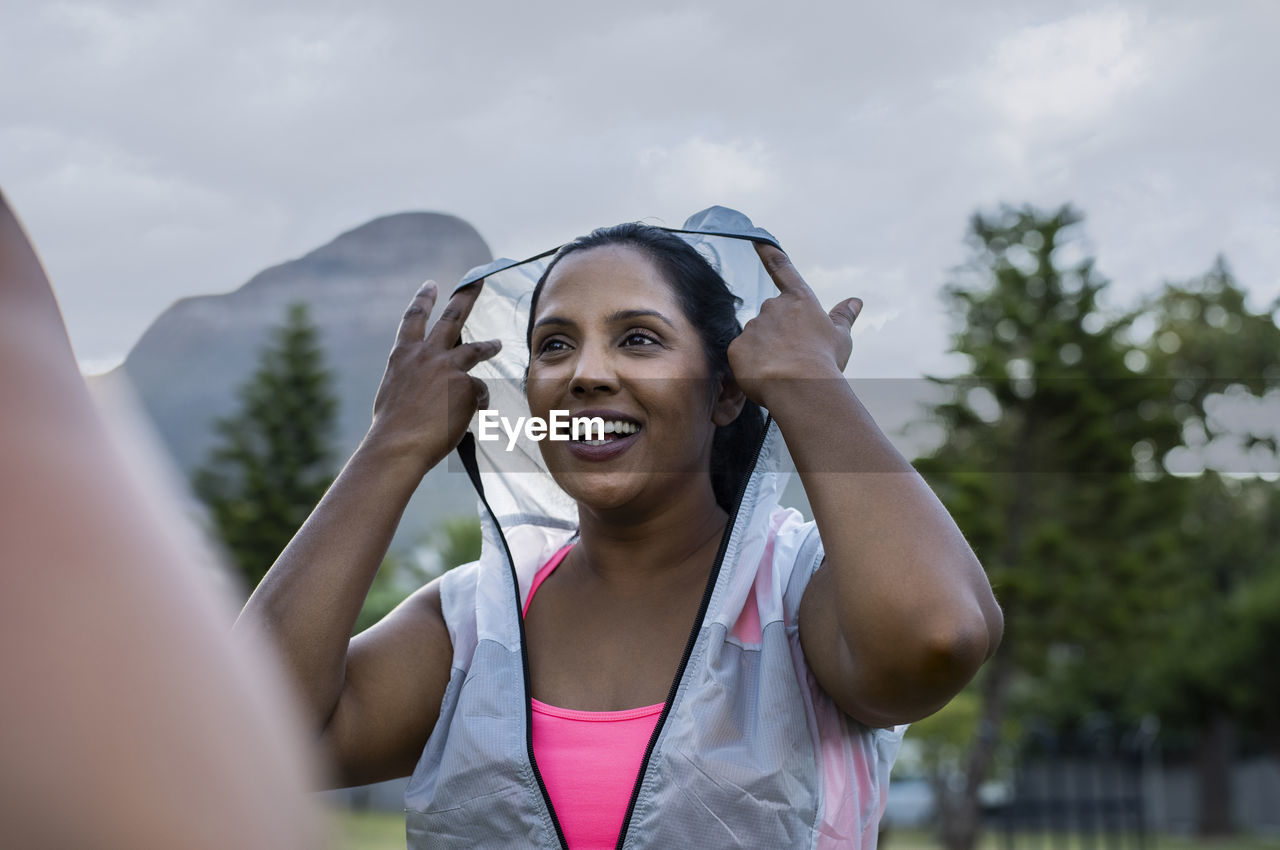 Low angle view of happy woman wearing hooded jacket while standing against sky in park