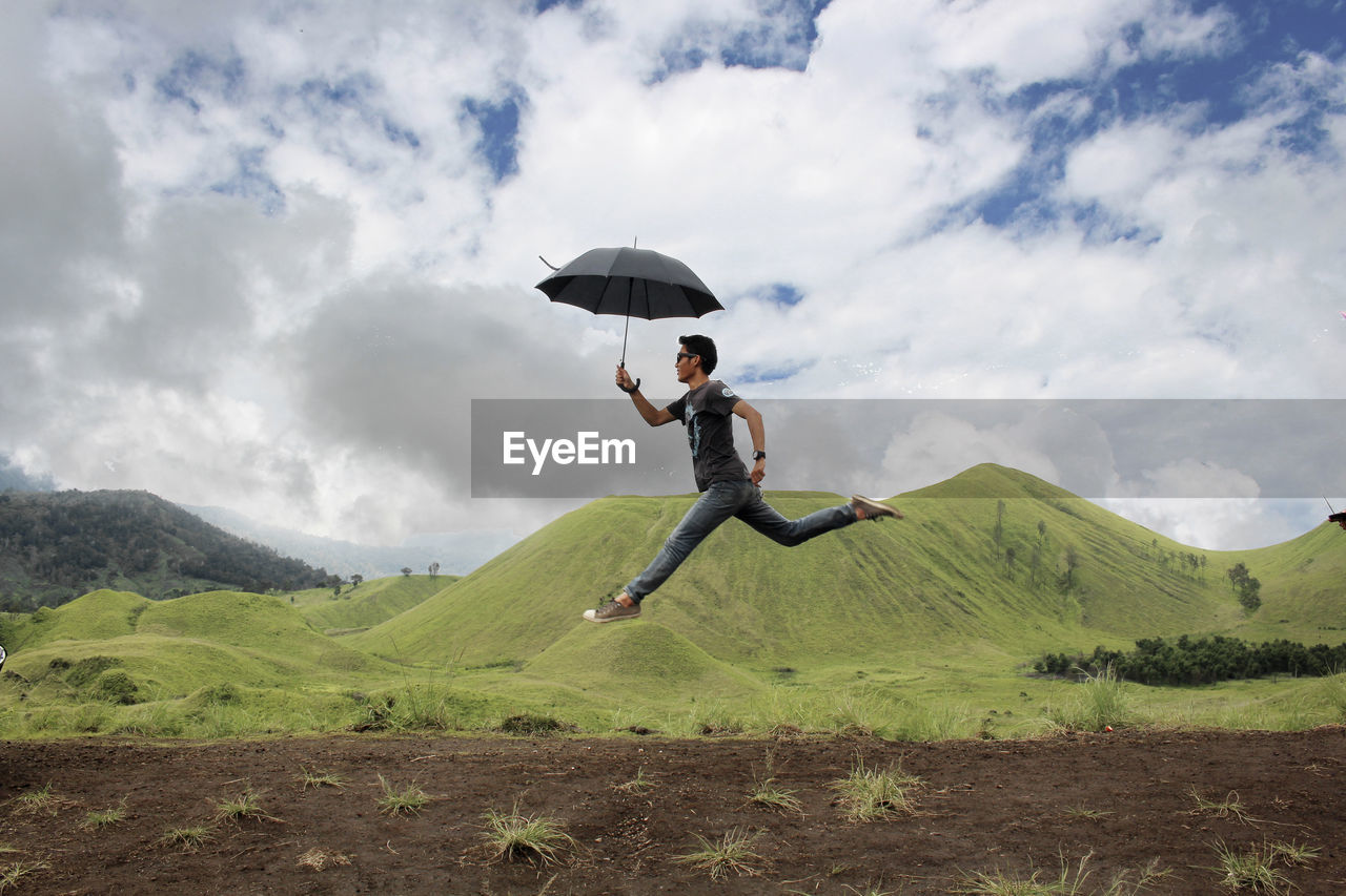 Side view of young man with umbrella jumping by on mountain against cloudy sky