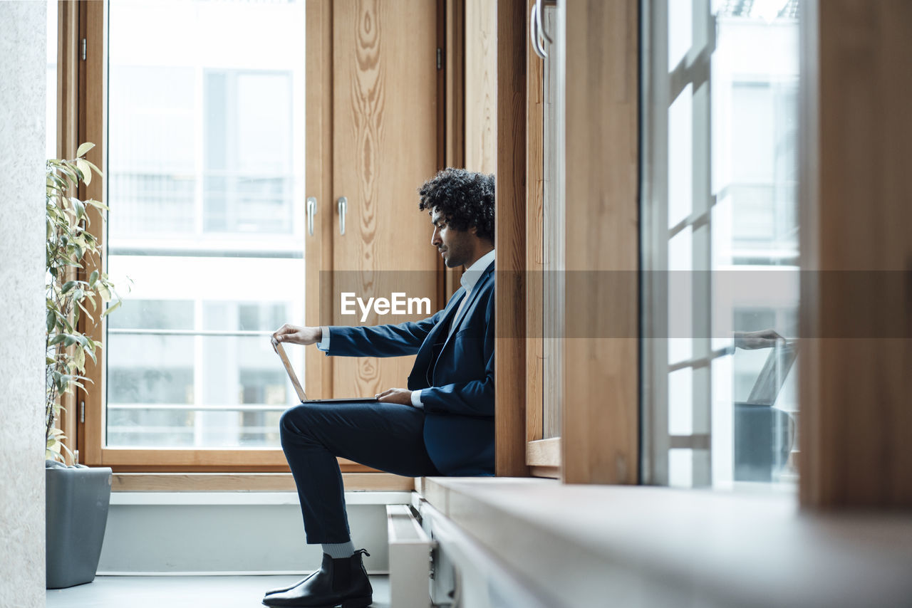 Young male entrepreneur using laptop while sitting against window at workplace