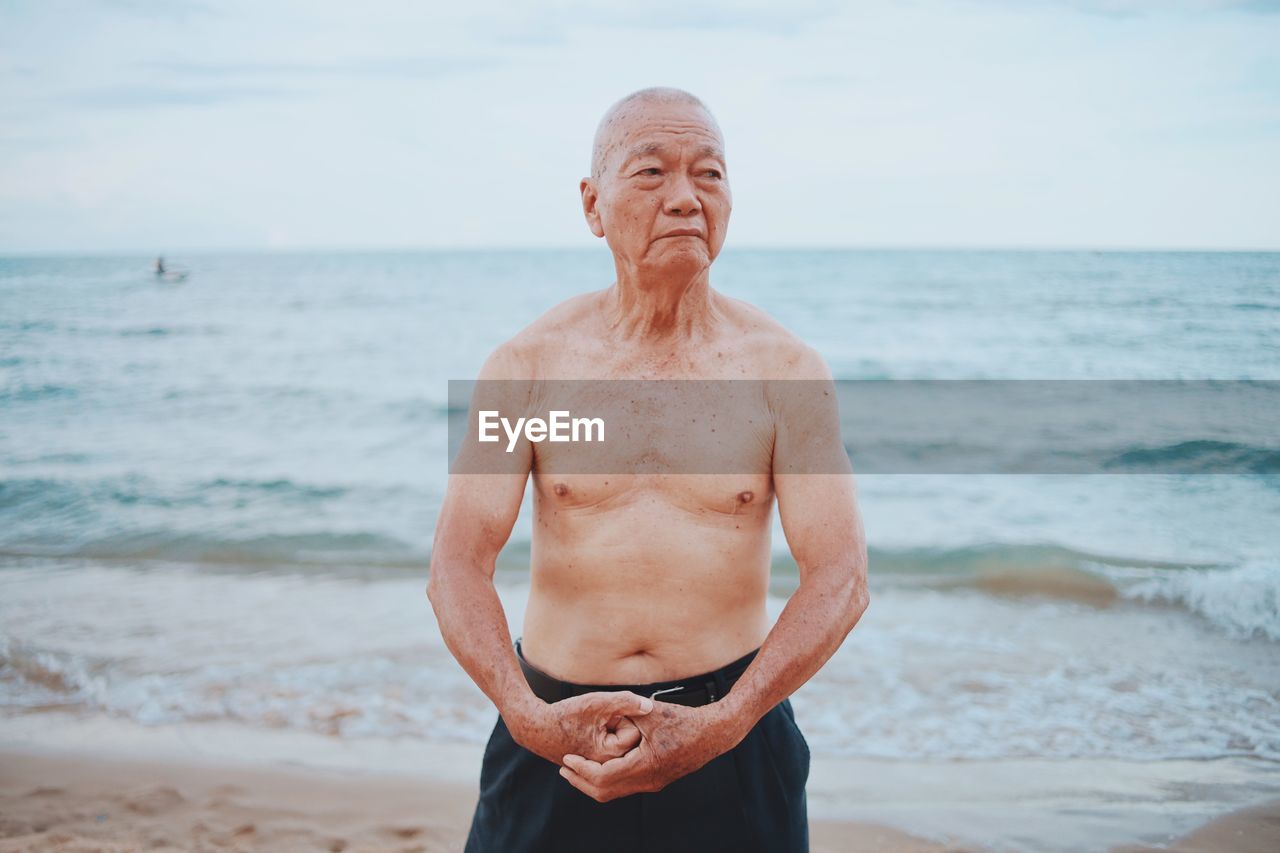 Shirtless senior man showing muscles while standing at beach against sky during sunset
