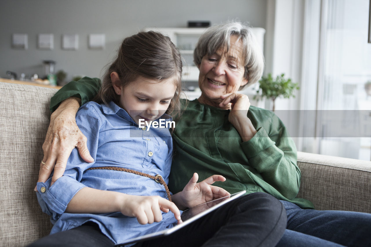 Grandmother and her granddaughter sitting together on the couch with digital tablet