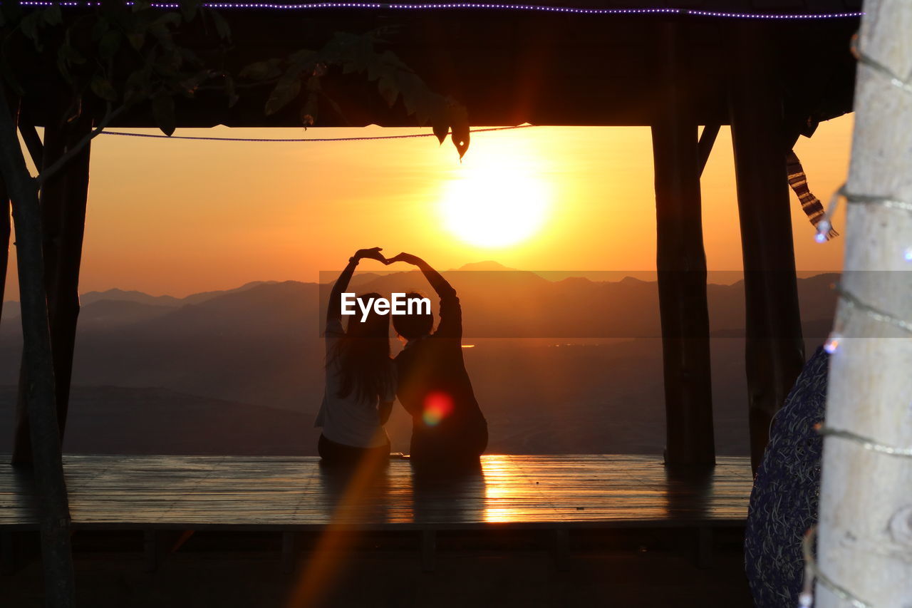 Rear view of friends making heart shape against sky during sunset