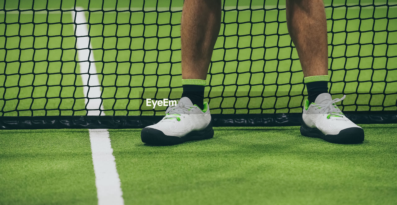 Closeup view of male legs with shorts in a padel tennis court standing near the net. foot on green