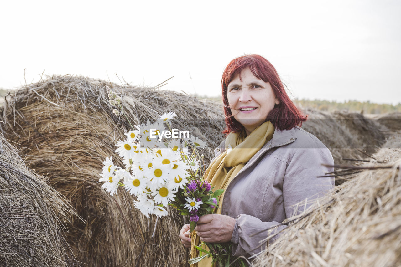 Senior woman holding bunch of chamomile flowers while standing amidst hay bales