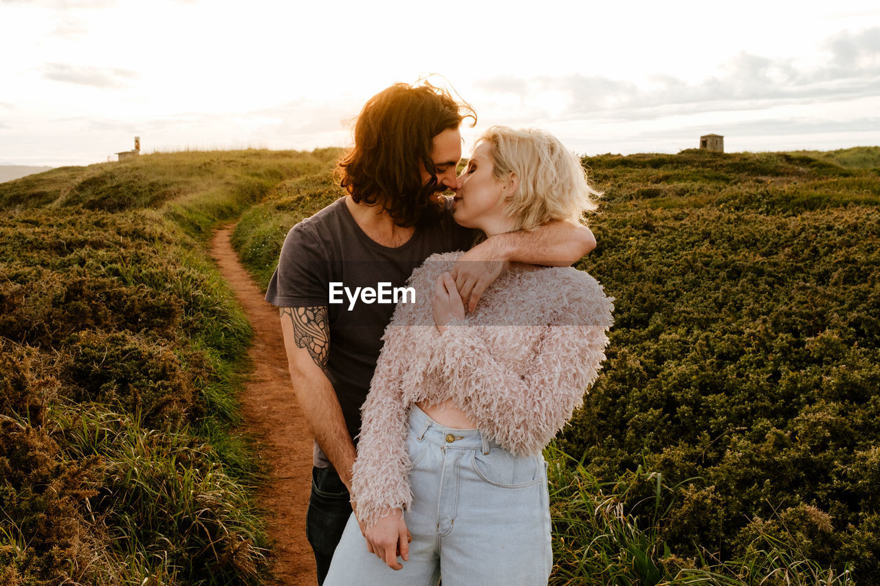 Bearded man embracing and kissing blond girlfriend while standing on grassy cliff at sunset in aviles, spain