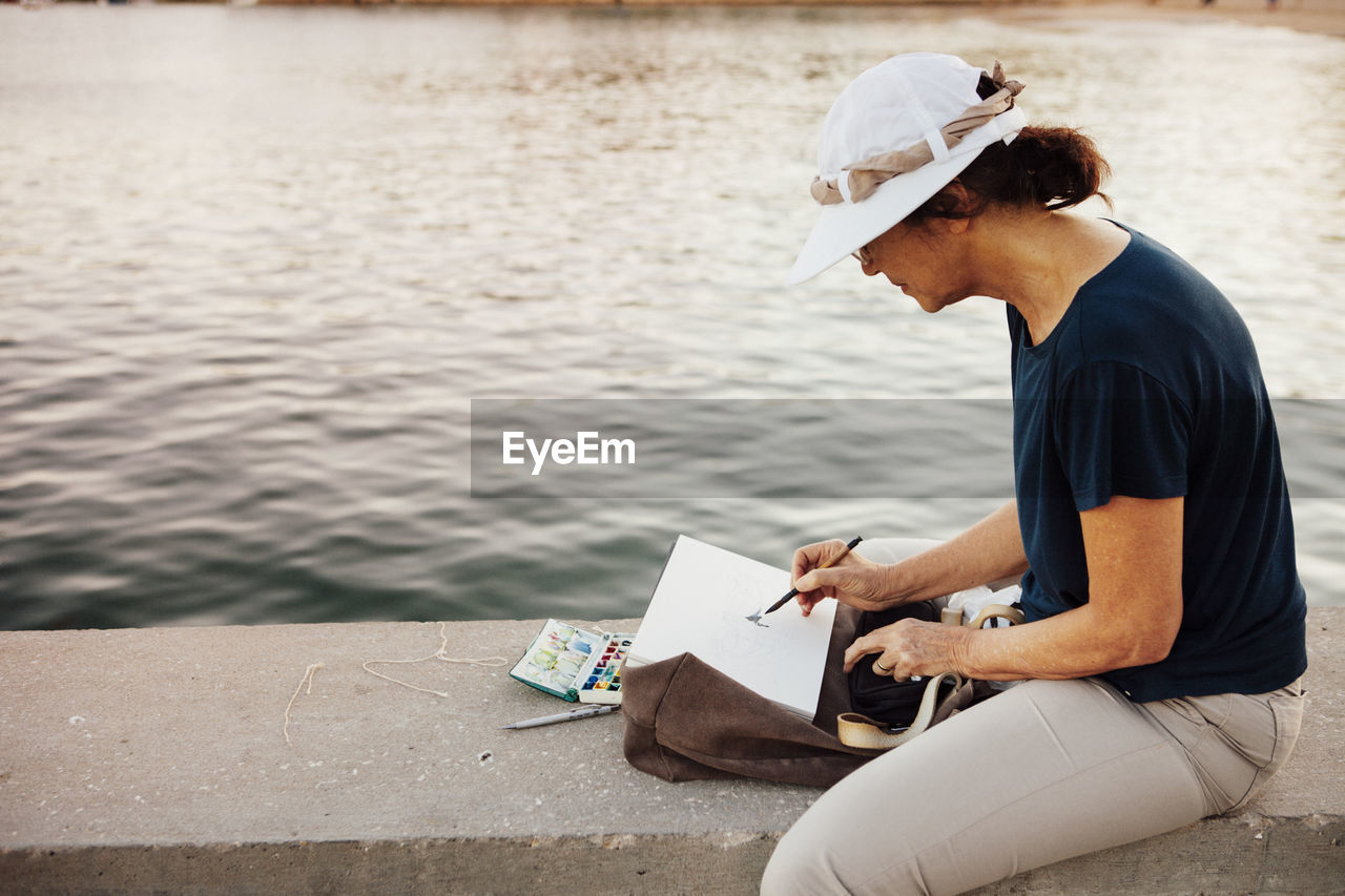 Side view of senior woman painting while sitting on retaining wall by sea