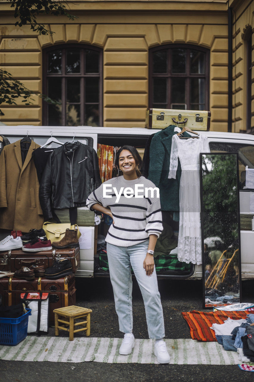 Portrait of happy female owner standing with hand on hip in front of van during second hand sale at flea market
