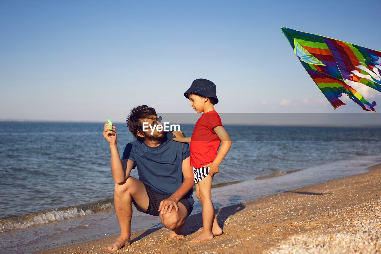 Father and son are standing on a sandy beach by the sea and launch a toy striped kite in the summer