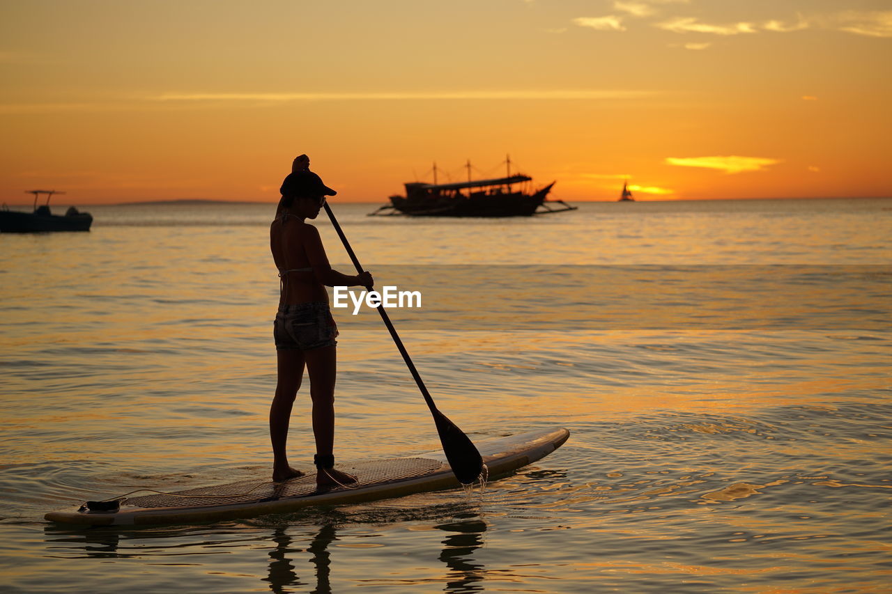 Silhouette woman paddleboarding in sea against orange sky