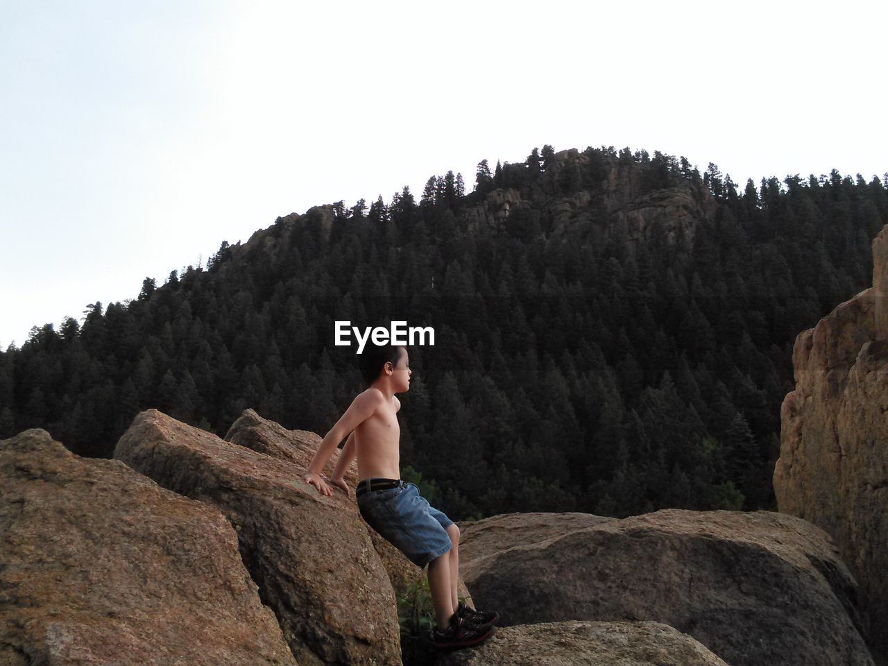 Full length of shirtless boy leaning on rock against mountain