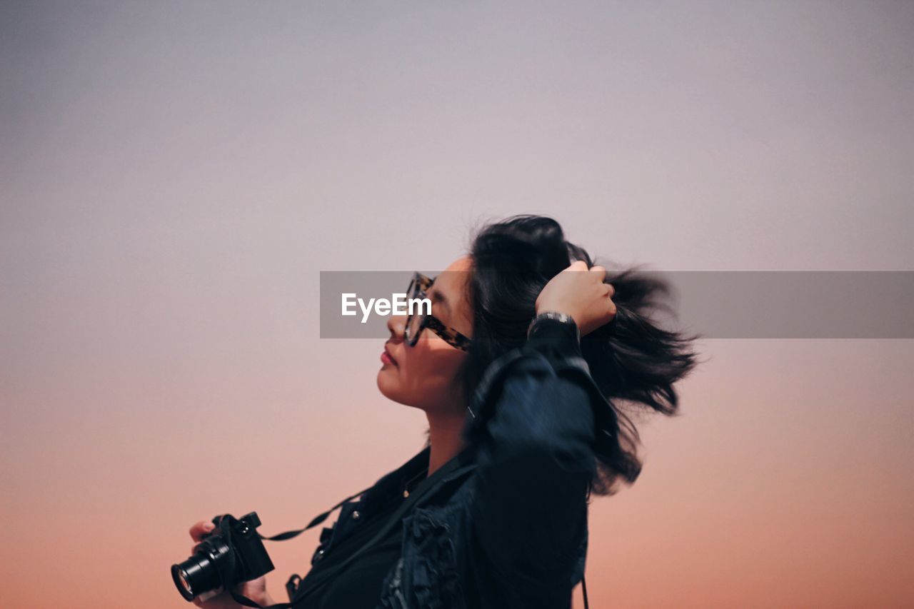 Young woman with hand in hair holding camera while standing against sky during sunset