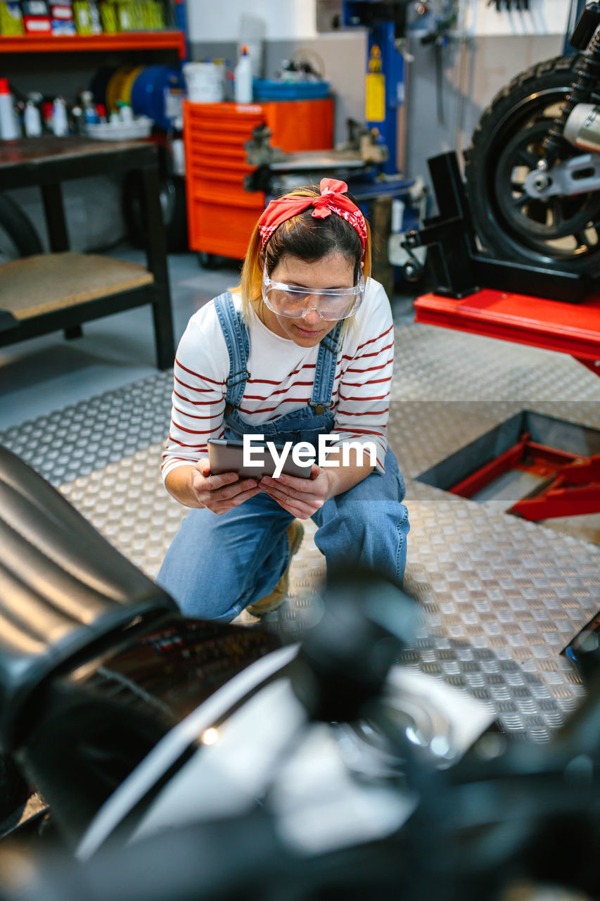 Concentrated female mechanic with security glasses holding digital tablet while review motorcycle