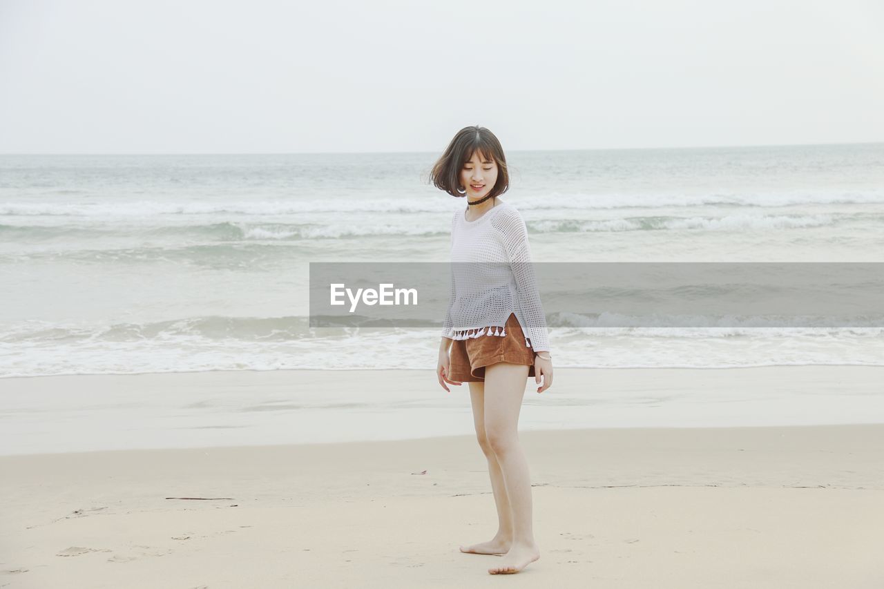 Side view of young woman standing at beach against clear sky