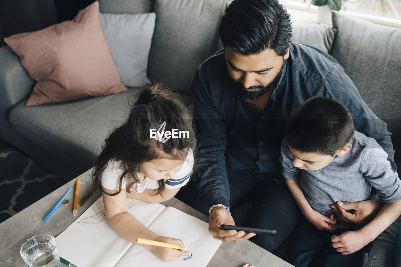 High angle view of father showing mobile phone to son while daughter drawing in book at home