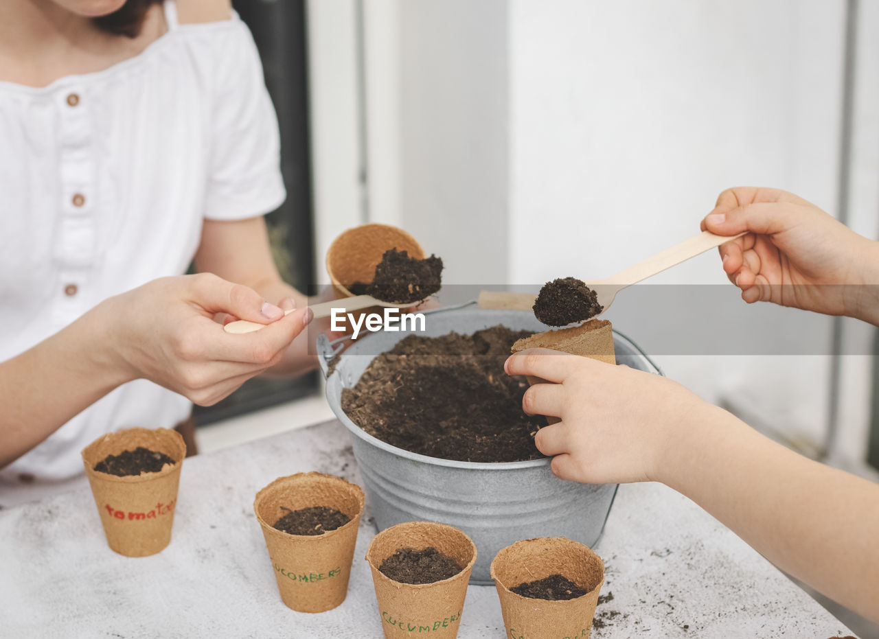 Two girls pour earth with wooden spoons from a zinc bucket into cardboard cups.