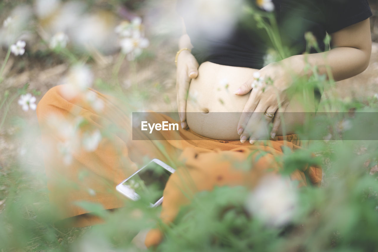 Midsection of pregnant woman with hands on stomach sitting amidst plants on field