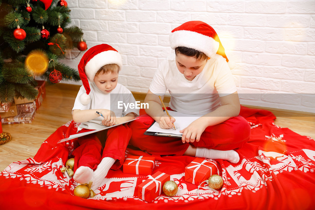 Cute sibling writing on envelope while sitting by christmas tree