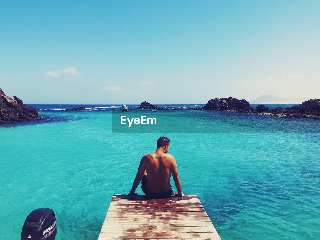 MAN SITTING AT SEA SHORE AGAINST SKY