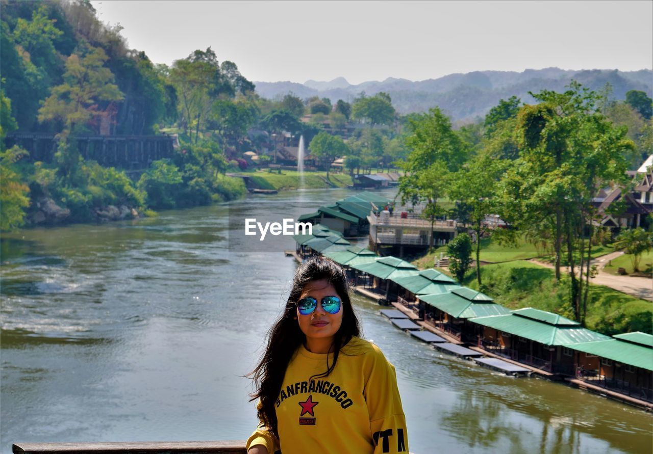 PORTRAIT OF BEAUTIFUL YOUNG WOMAN STANDING BY TREES AT RIVERBANK