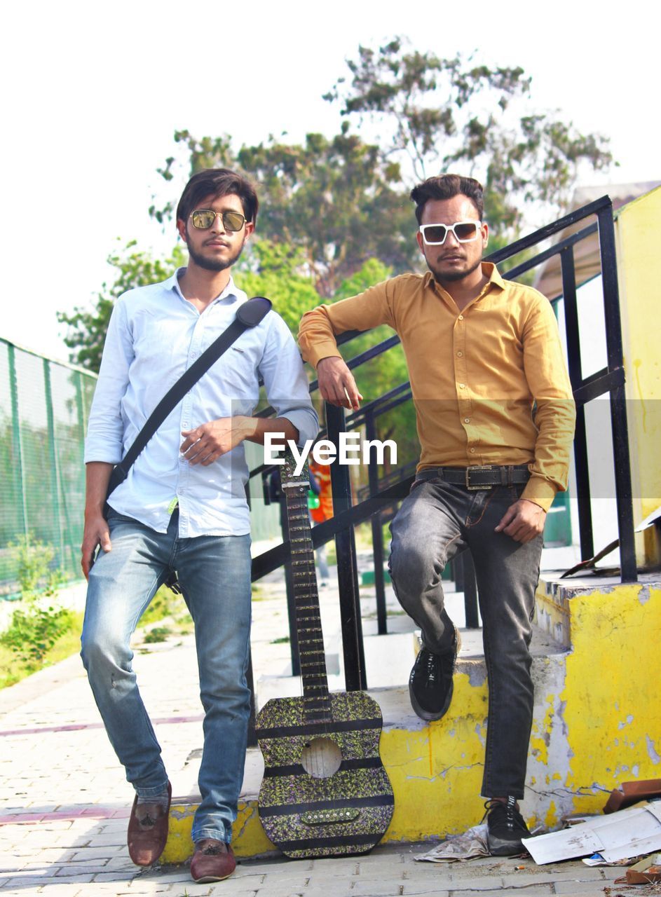 men, adult, two people, full length, young adult, casual clothing, togetherness, fashion, smiling, day, friendship, sunglasses, happiness, architecture, portrait, city, jeans, standing, glasses, emotion, front view, looking at camera, outdoors, lifestyles, leisure activity, clothing, footwear, transportation, cool attitude, person, nature, tree, city life