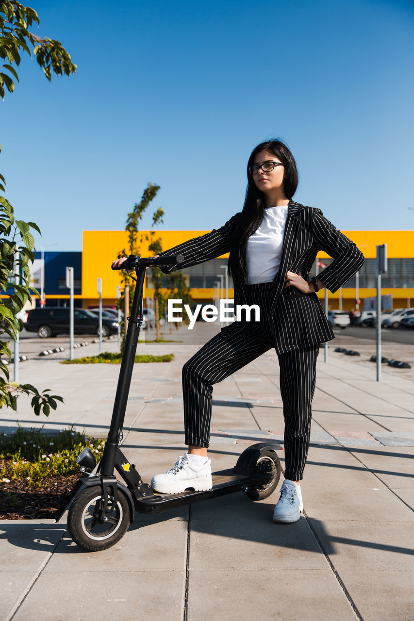 Woman with push scooter standing on footpath against clear blue sky