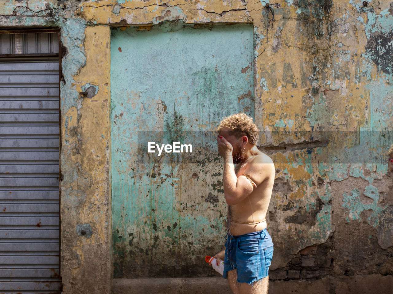 Full length of shirtless man standing against wall with hand to face.