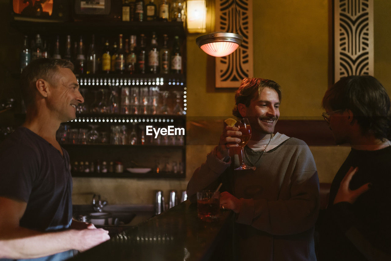 Male bartender looking at gay couple enjoying drinks at bar