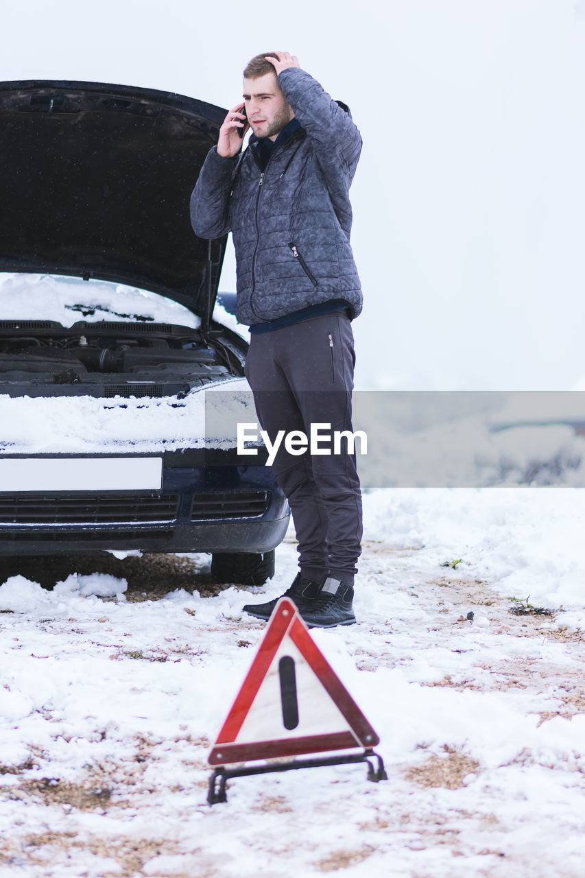 Young man talking over mobile phone while standing by car on snow covered field against sky