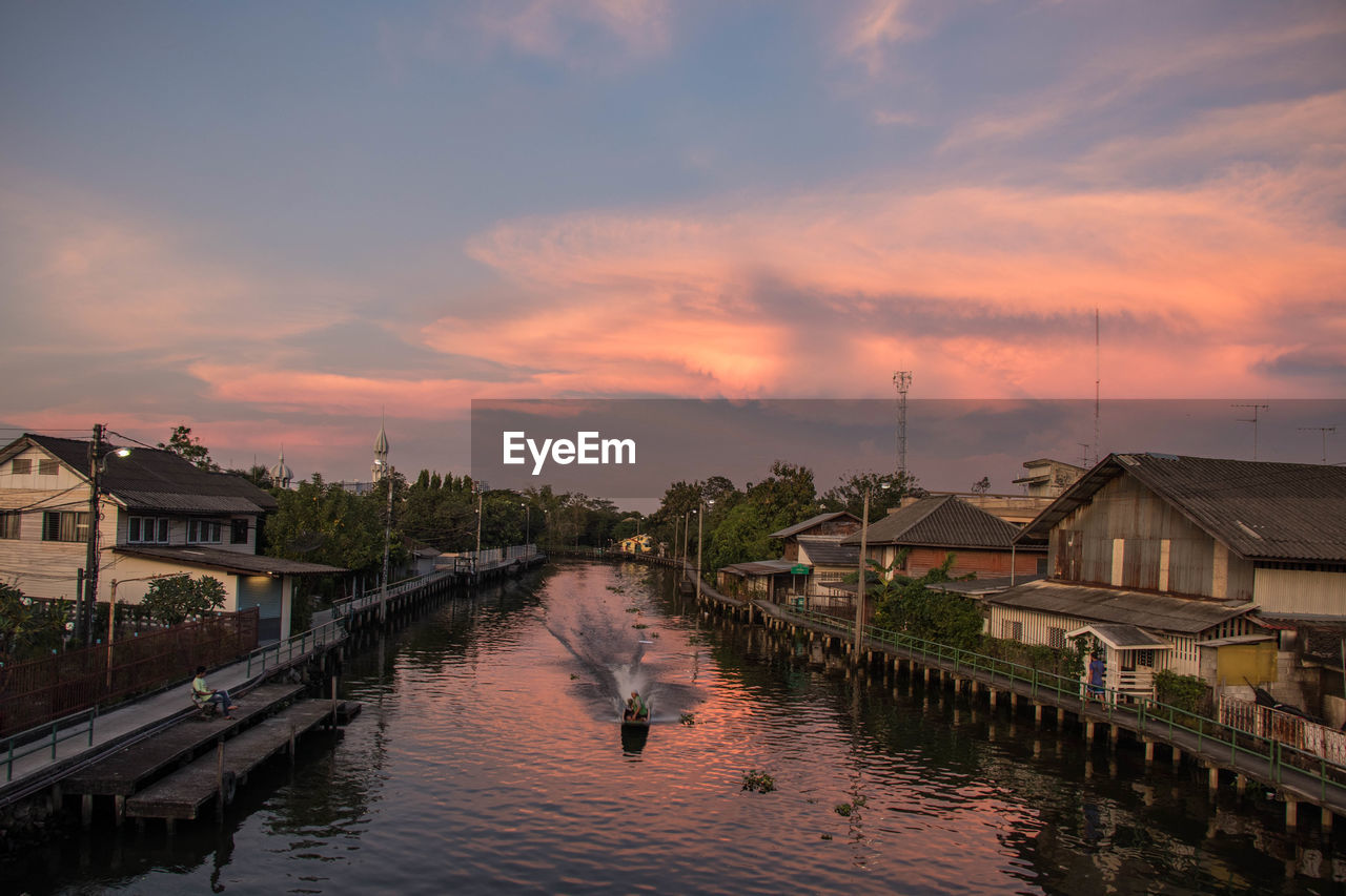 RIVER AMIDST BUILDINGS AGAINST SKY AT SUNSET