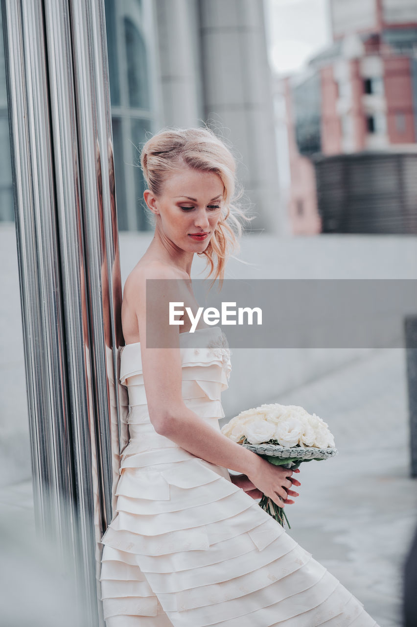 Beautiful bride with bouquet against wall in city