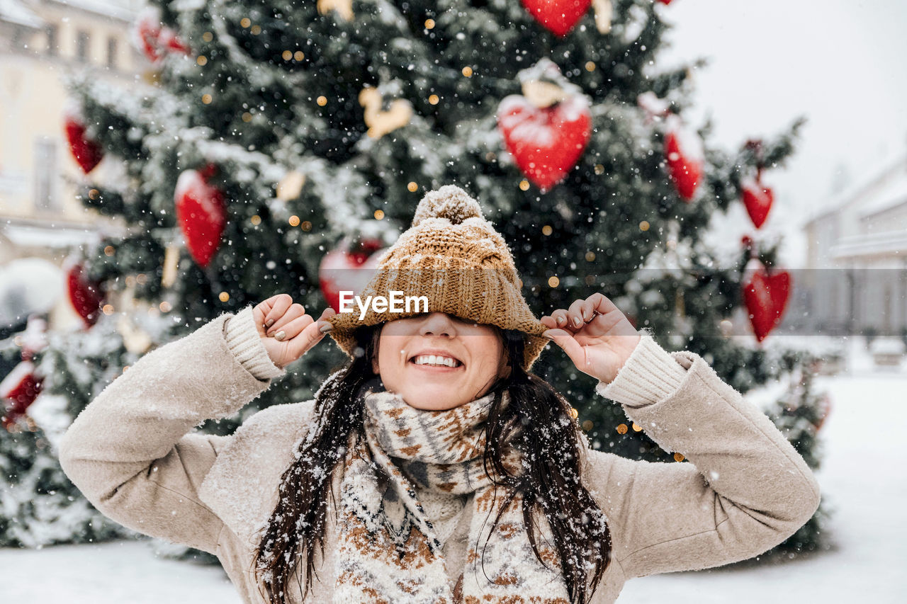 Portriat of young woman pulling her hat over her eyes, happy, smiling, winter, christmas, snowing.