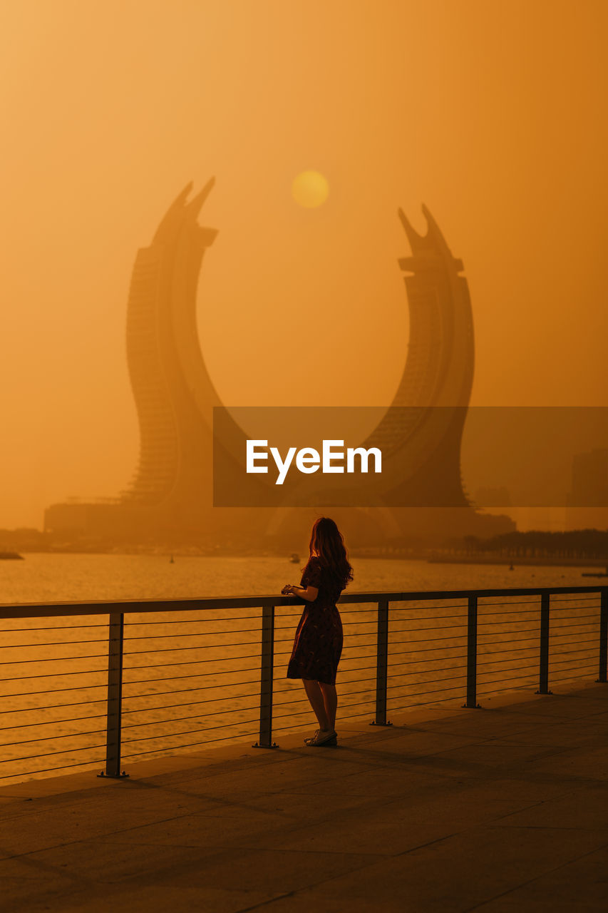 Back view of young female tourist with short dark hair in casual clothes standing on embankment and admiring creative moon shaped building against scenic sunset sky in doha