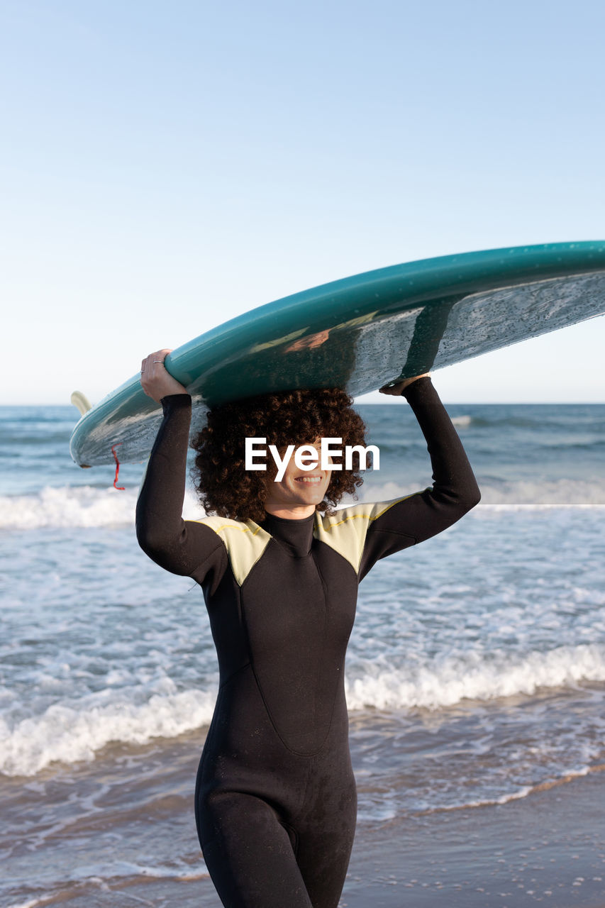 Side view of young happy female surfer in wetsuit with surfboard standing holding surfboard above head looking away on seashore washed by waving sea