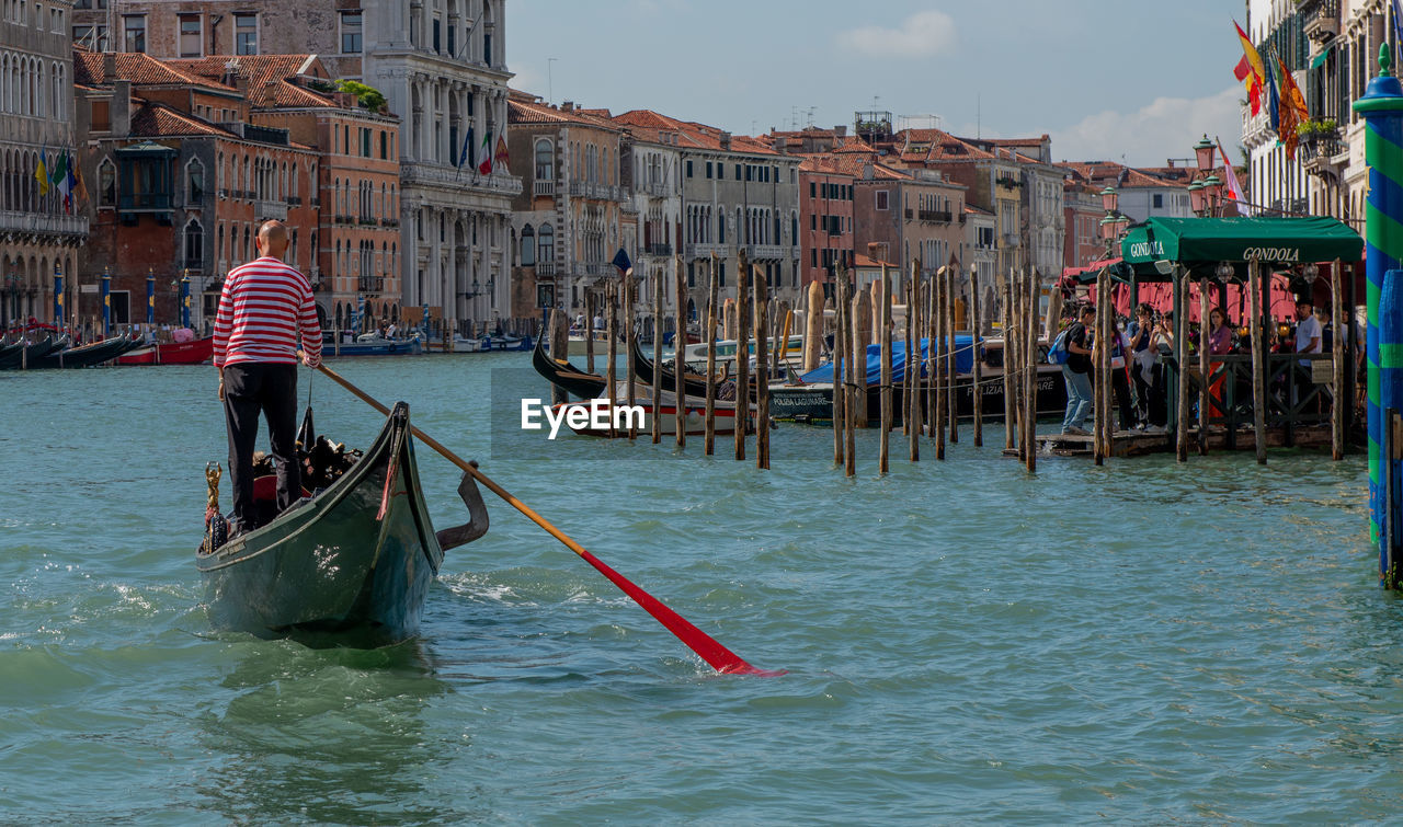 gondola, nautical vessel, water, transportation, canal, mode of transportation, travel destinations, gondolier, travel, architecture, tourism, building exterior, boat, nature, vehicle, built structure, city, boating, trip, vacation, holiday, sea, tourist, lagoon, men, occupation, watercraft, outdoors, waterfront, sky, tradition, rowing, adult, oar, wooden post, day, group of people, waterway, bridge, romance, lifestyles, rear view
