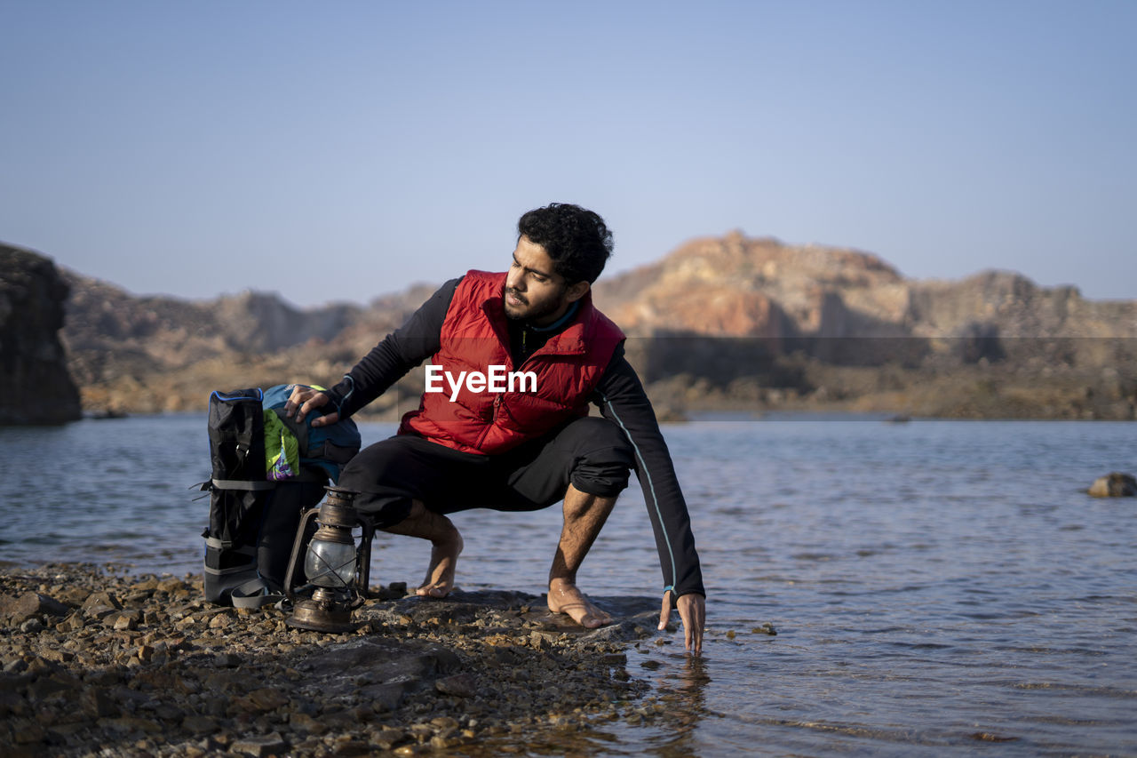 Young indian traveler getting ready for camping in the mountains, sitting near a lake.