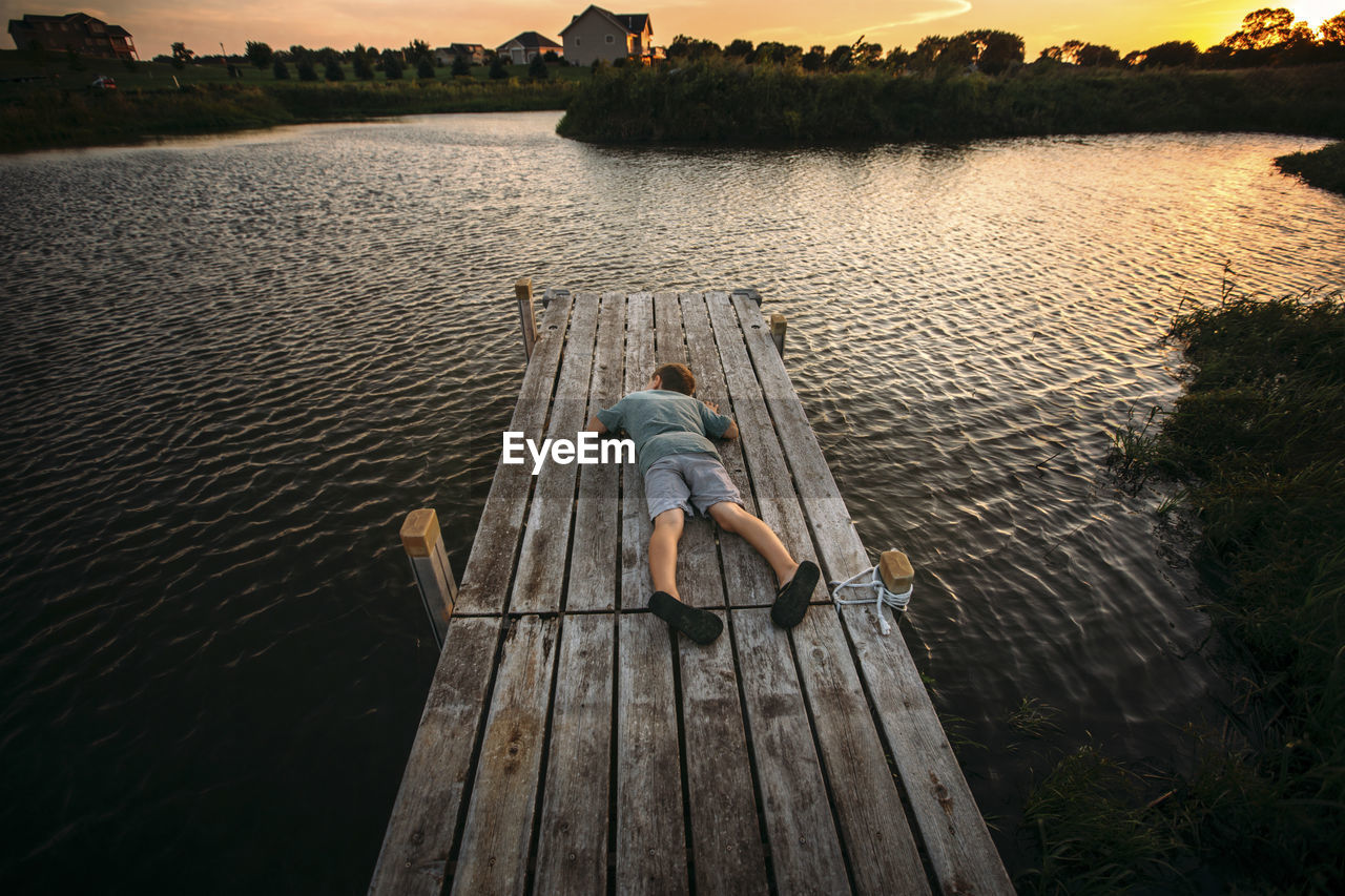 High angle view of boy lying down on jetty against sky during sunset