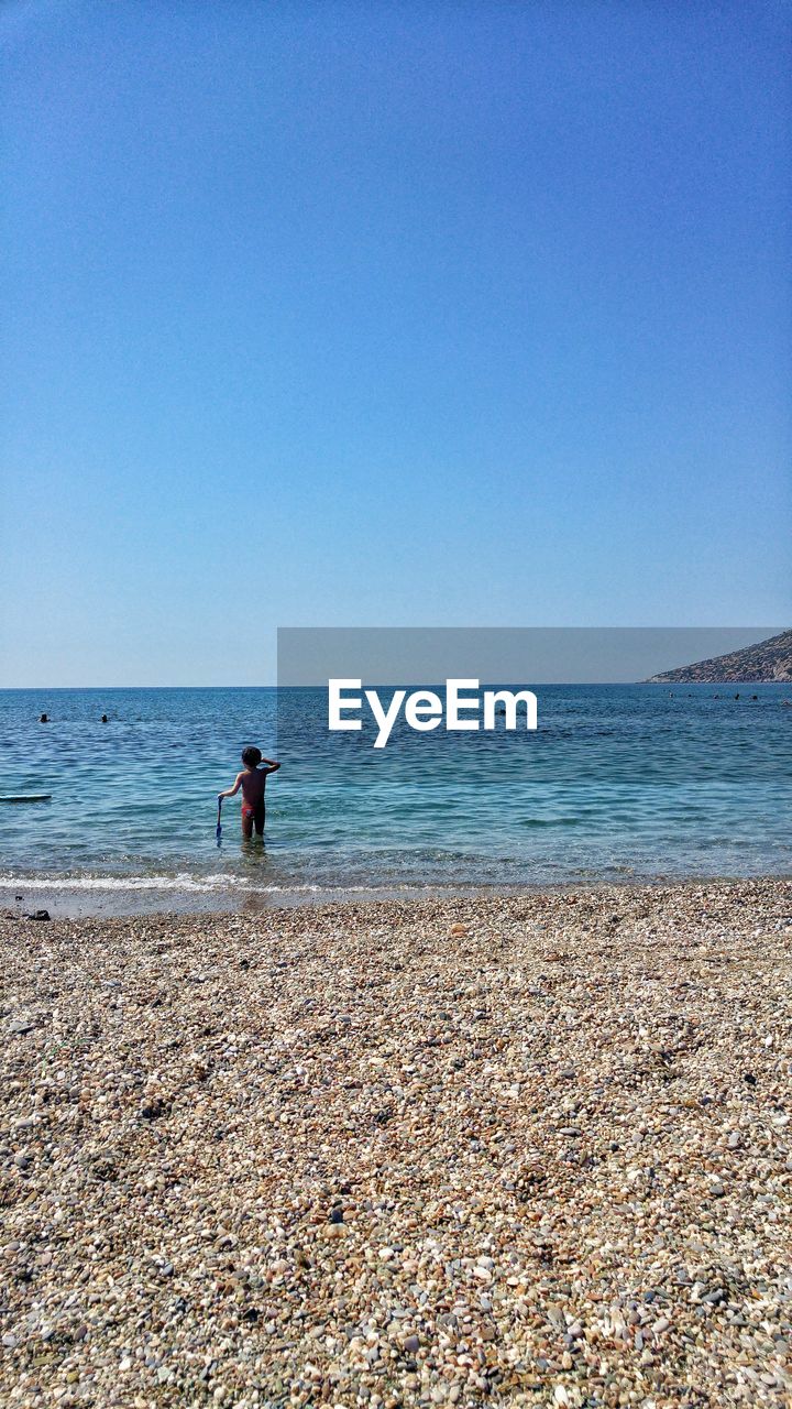 Rear view of boy standing on shore at beach against clear blue sky