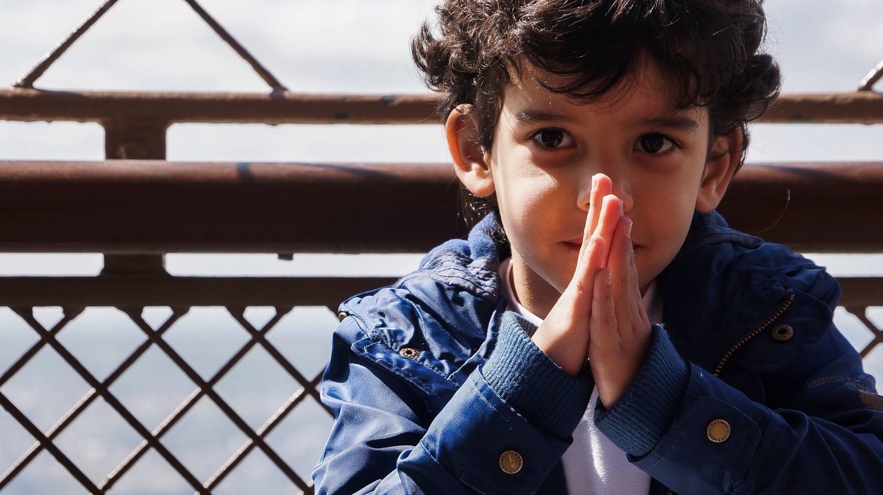 Close-up portrait of boy with hands clasped against fence