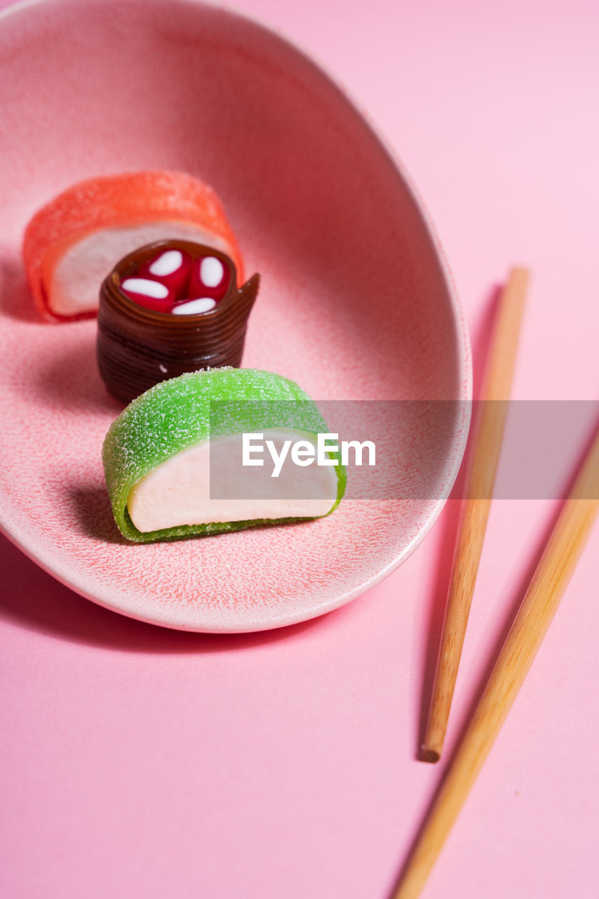 Appetizing colorful sweet sushi served on ceramic plate with wooden chopsticks placed on pink background