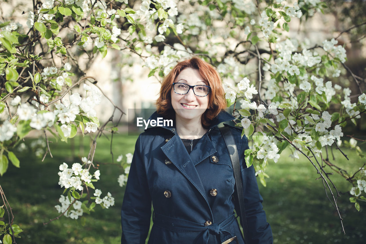 Confident middle aged armenian woman in a blue trench coat and glasses under the blooming tree