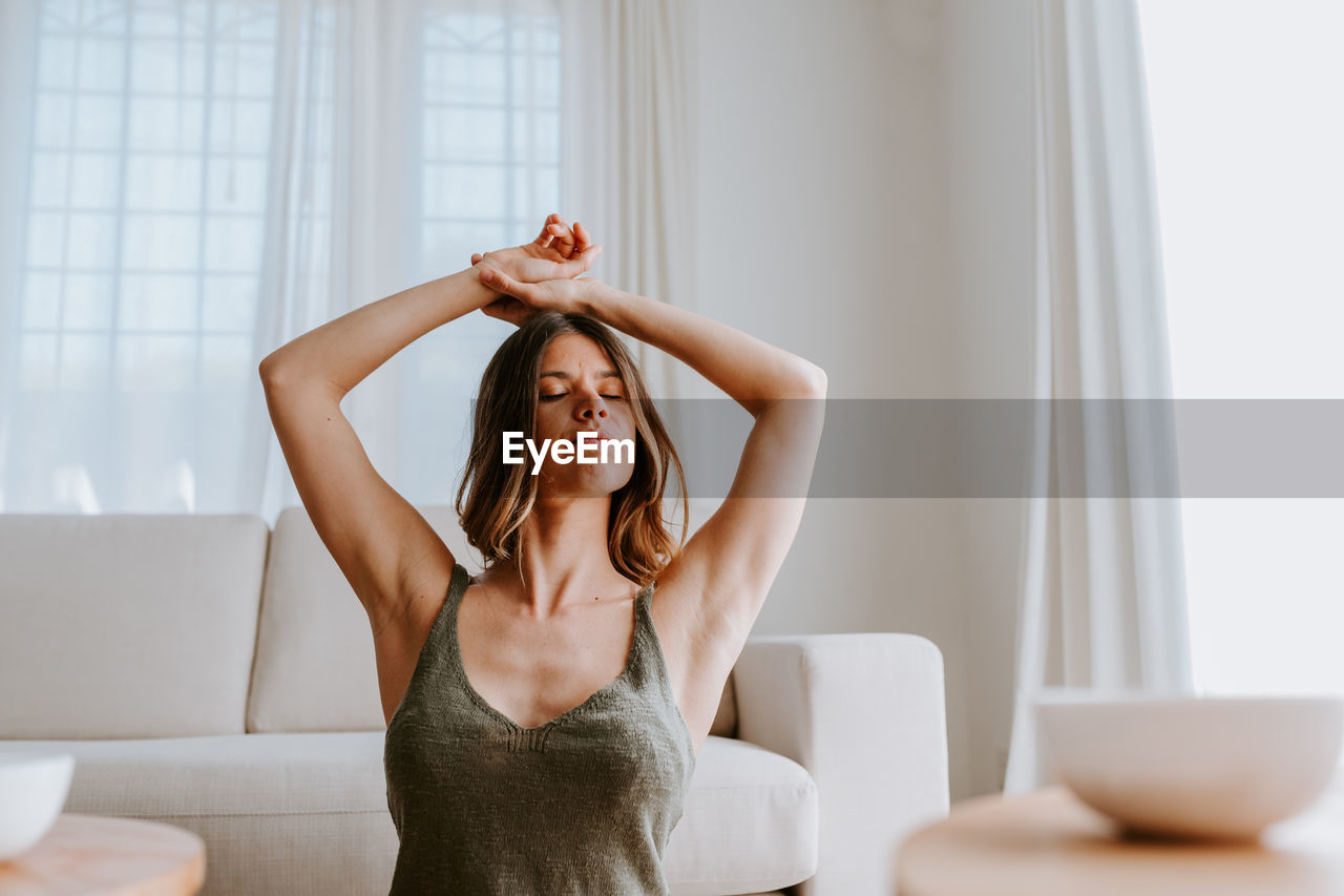 Tranquil female stretching arms while sitting on floor at home and enjoying calm morning with eyes closed