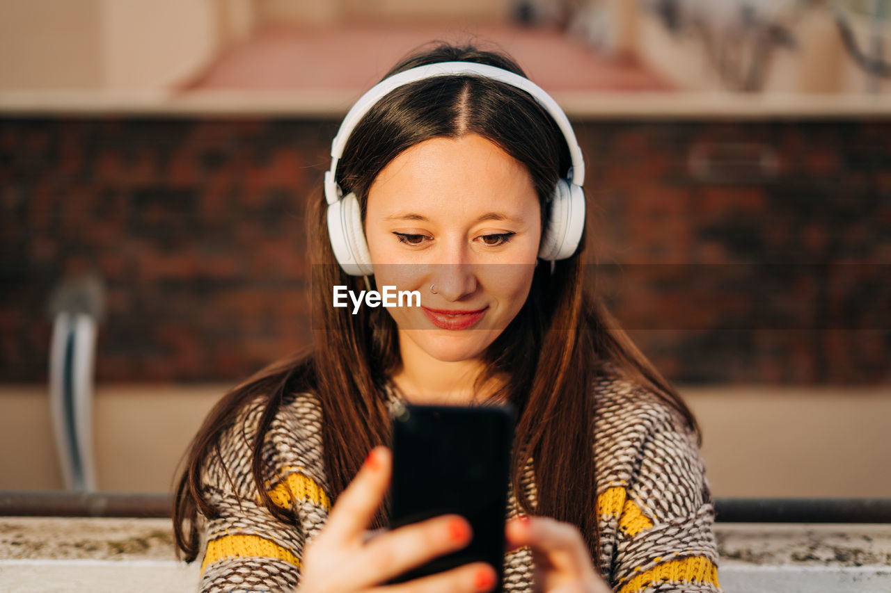 Smiling woman wearing headphones and using phone at home 