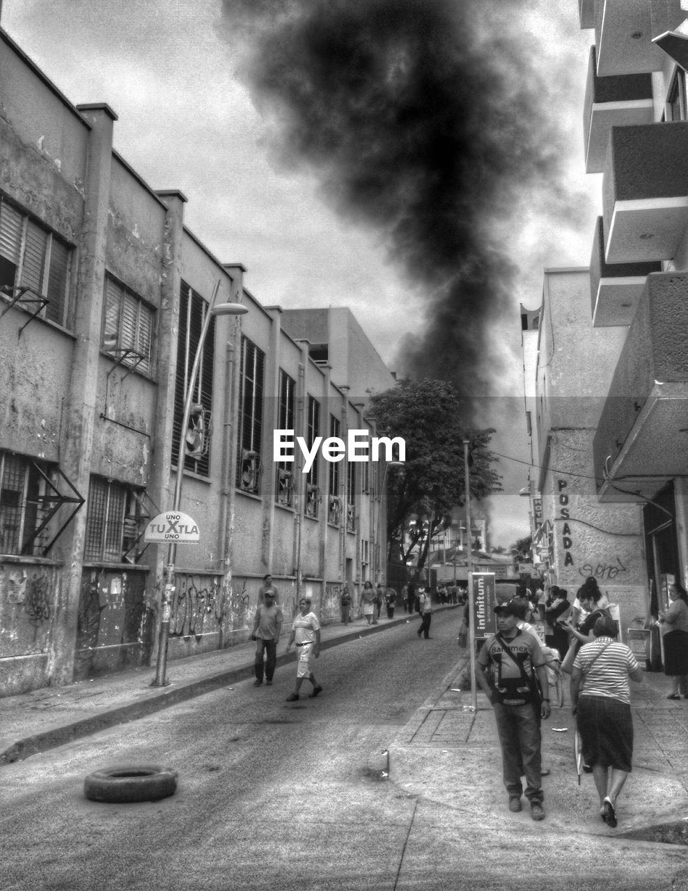 People walking on city street by smoke against sky