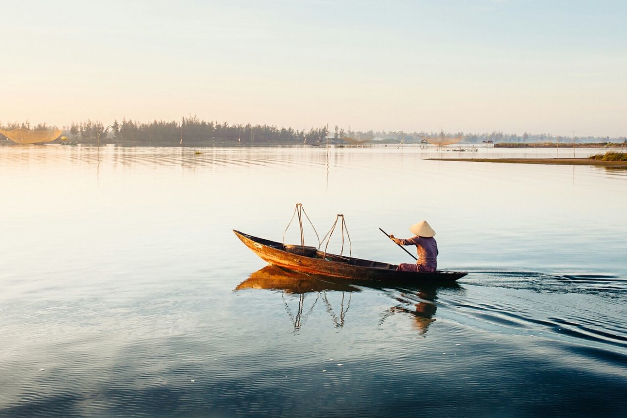 Rear view of fisherman wearing asian style conical hat on boat at lake against sky