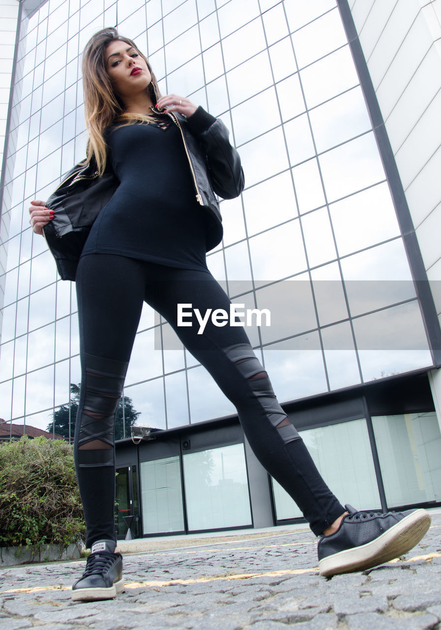 Low angle portrait of young woman standing on footpath against building