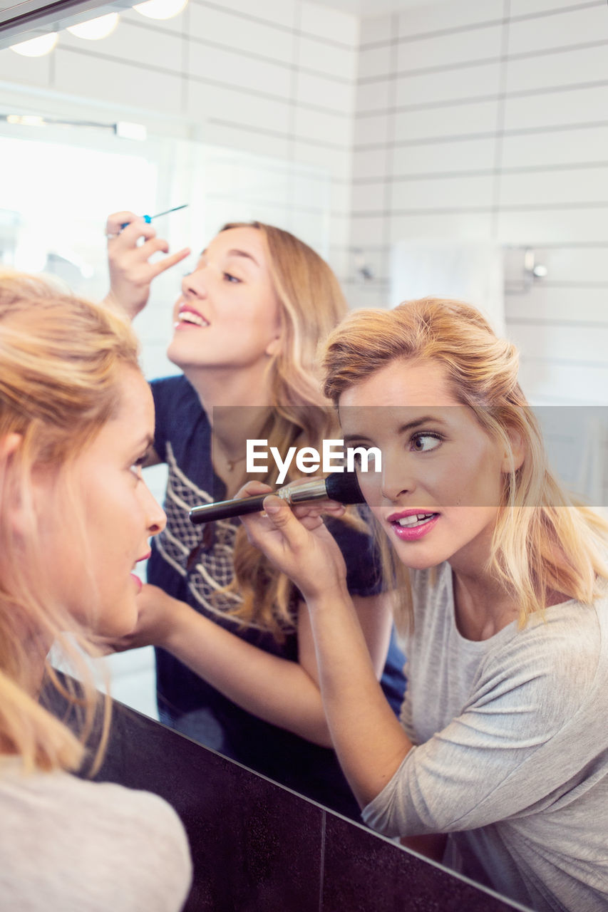 Young female friends applying makeup in mirror