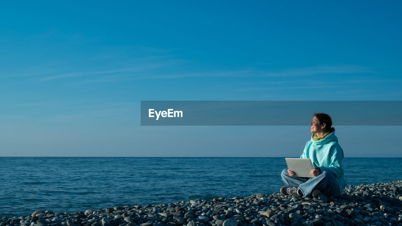 Caucasian woman working on a laptop on a pebbly seashore.